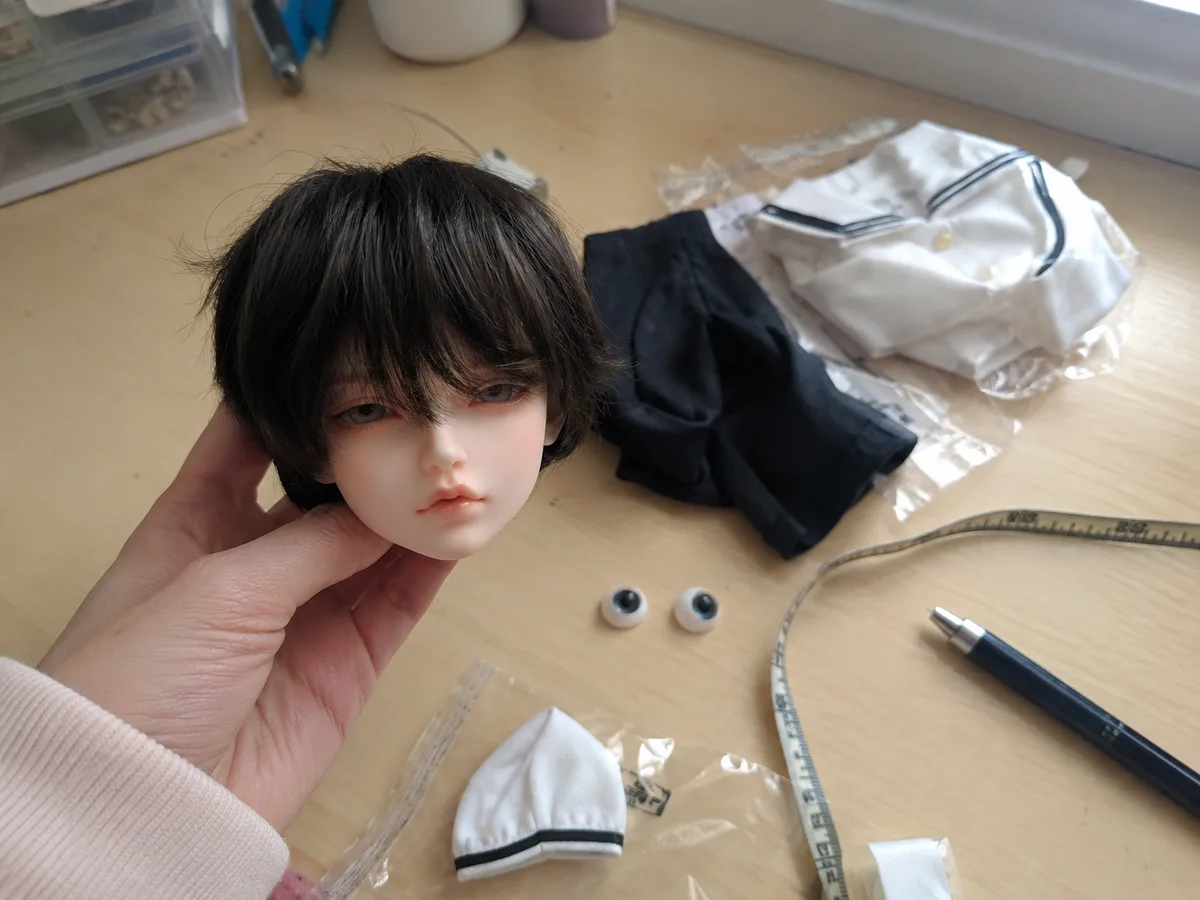 Fit check in hand: A buyer holds the doll head while checking eyes, wig, clothes, and measurement tape on a home desk.